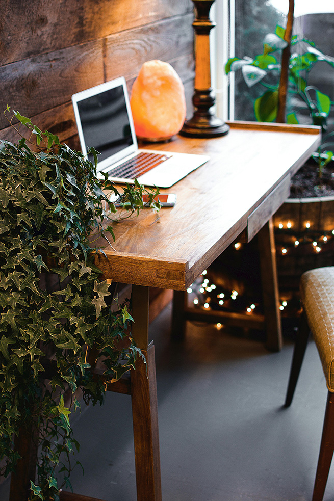 A cozy wooden desk holds an open laptop, glowing salt lamp, and small plant, with trailing greenery, warm string lights, and a leafy window view.