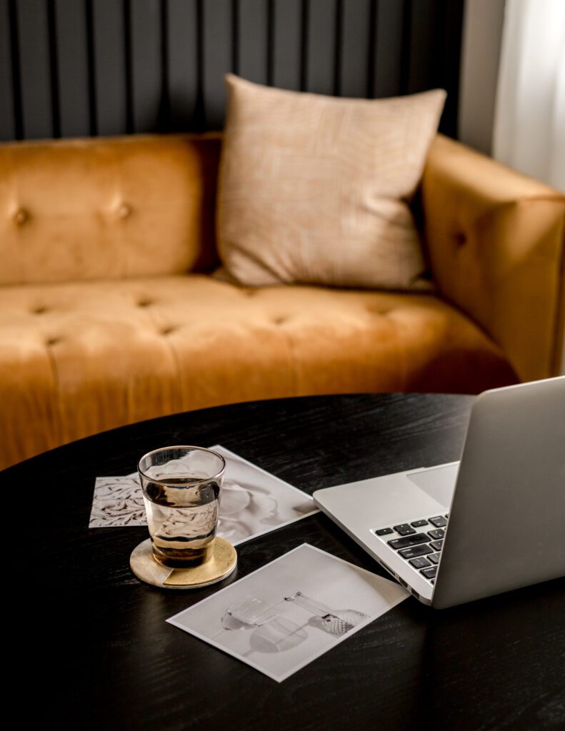 Laptop on a coffee table in front of a brown leather couch, representing a cozy and organized workspace for managing a wellness business with Dubsado.