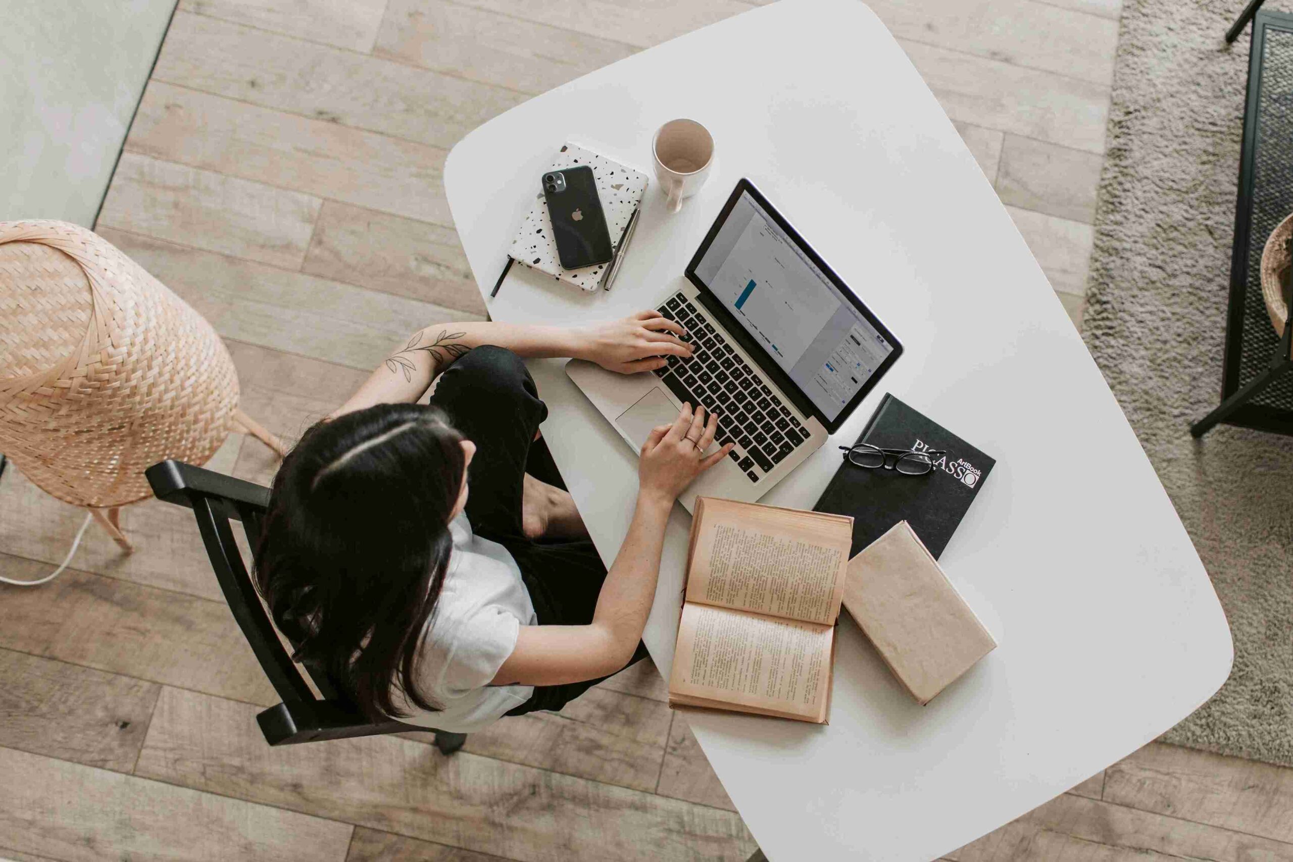 A woman seated at a table, working on a laptop surrounded by open books.