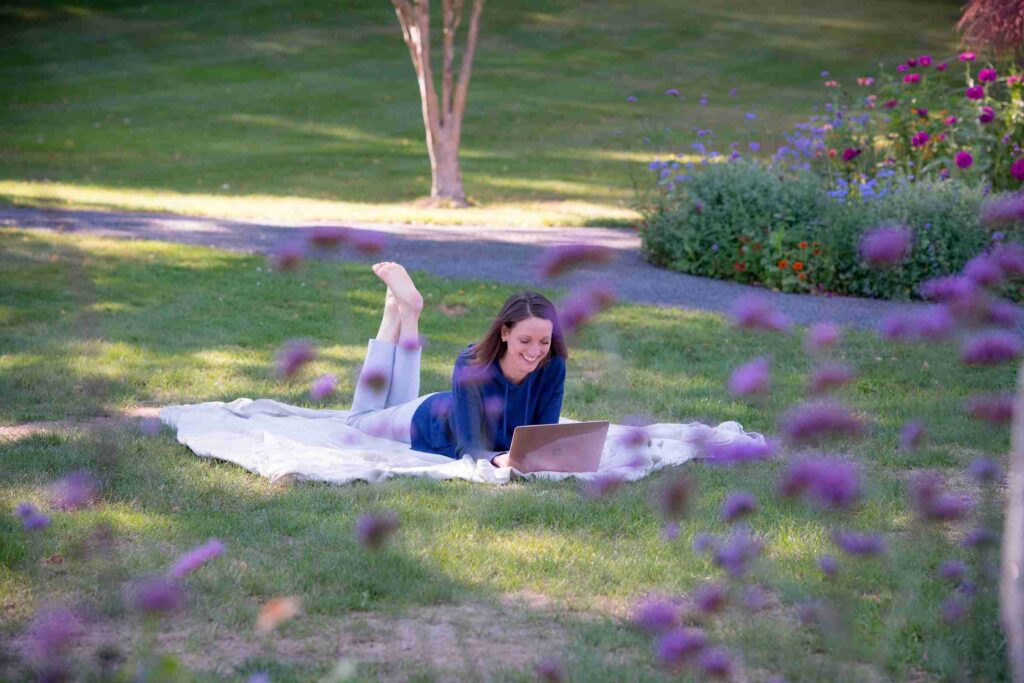 A woman relaxes on a blanket in the grass, focused on her laptop while enjoying the outdoors.