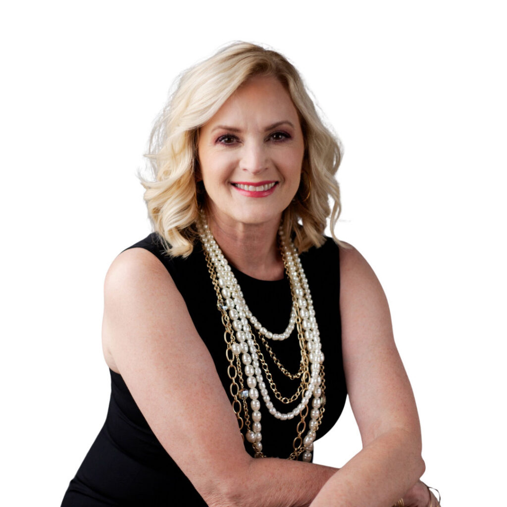 A smiling blonde woman poses against a white background in a black sleeveless top and layered pearl and gold necklaces, arms folded, with a warm, polished look.