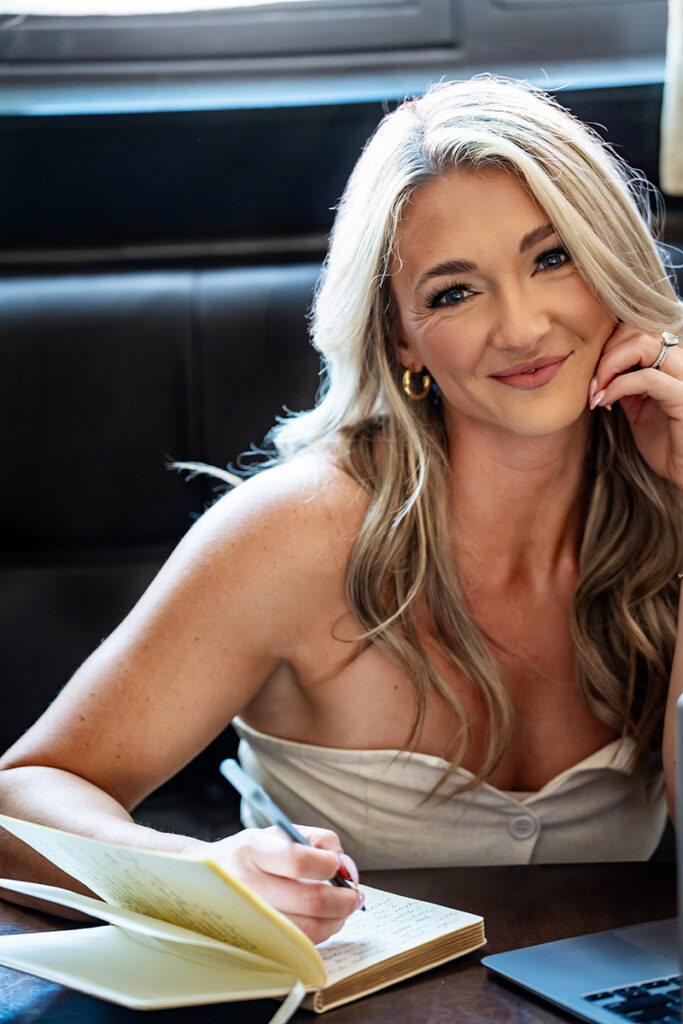 Smiling Zoe Schroeder in a cream top writes in an open notebook at a table, seated in a sleek black leather booth; warm, confident portrait.