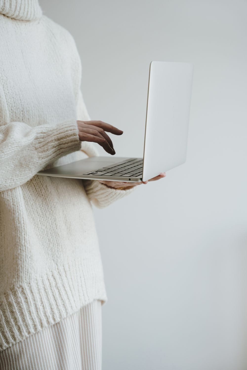 woman in a white sweater stands holding an open laptop.