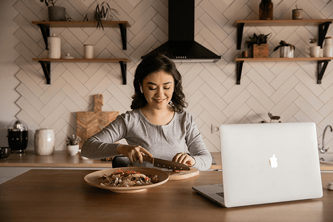 Nutritionist cutting vegetables in a cozy kitchen, smiling while preparing a meal. A laptop is open on the counter, surrounded by modern decor and plants.