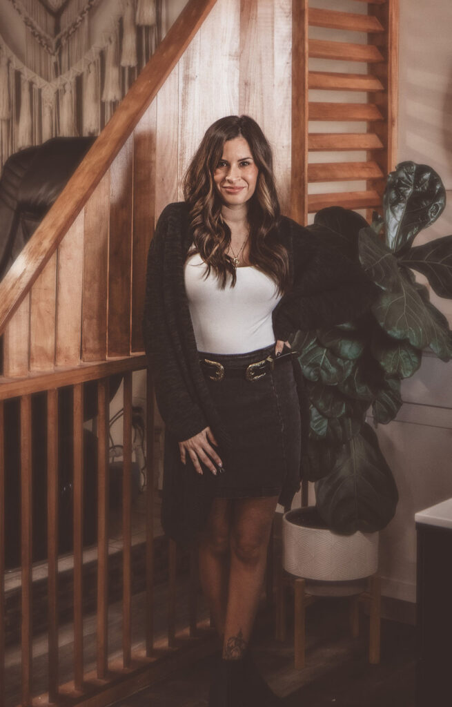 Andrea Duvall, salon owner, smiling with long dark hair poses beside a wooden staircase and leafy houseplant in a cozy home, wearing a black cardigan and white top.