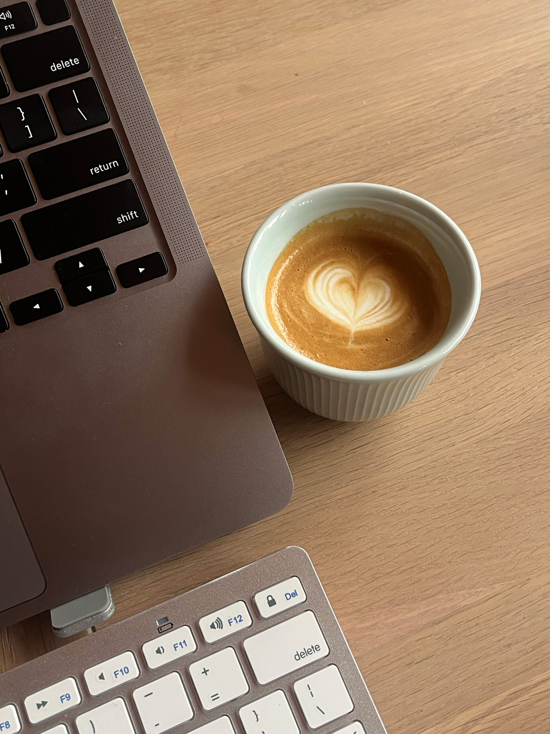 Close-up of a laptop and keyboard on a wooden desk, alongside a cappuccino in a white cup featuring heart-shaped latte art, creating a cozy work vibe.