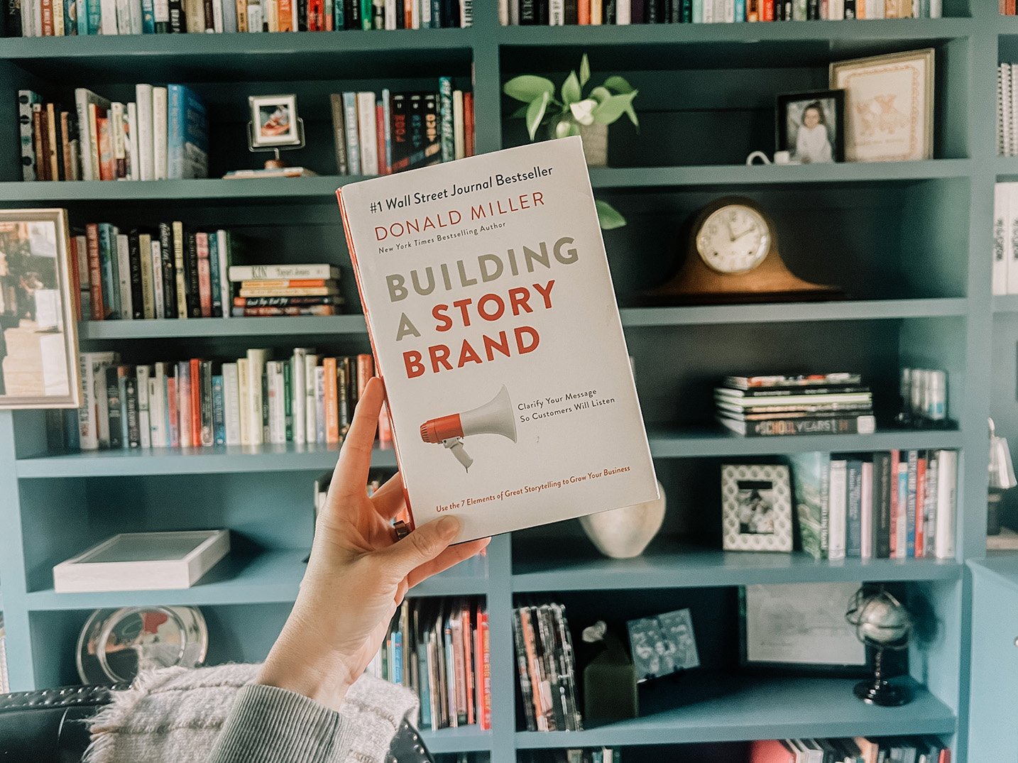 A hand holding the book "Building a Story Brand" by Donald Miller in front of a teal bookshelf filled with various books and decorative items, conveying a scholarly ambiance.