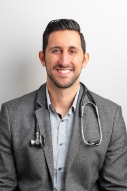 Smiling male doctor in a gray blazer and light shirt, wearing a stethoscope, poses against a plain white background with a warm, approachable expression.