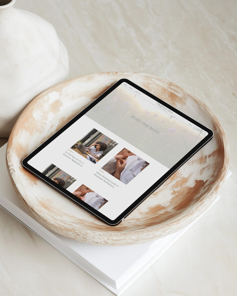 Tablet displaying a lifestyle webpage rests in a rustic ceramic bowl atop a white book, creating a calm, minimalist scene.