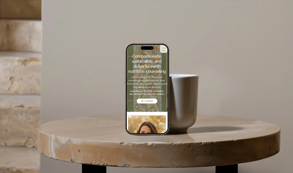 Upright smartphone showing a compassionate, sustainable nutrition counseling website on a rustic wooden table beside a mug in a calm, warm setting.