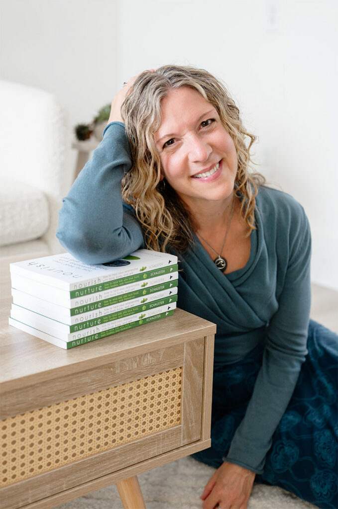 Heidi Schauster smiling in a teal top leans beside a wooden cane-front cabinet, resting her head on her hand near stacked books in a bright room.