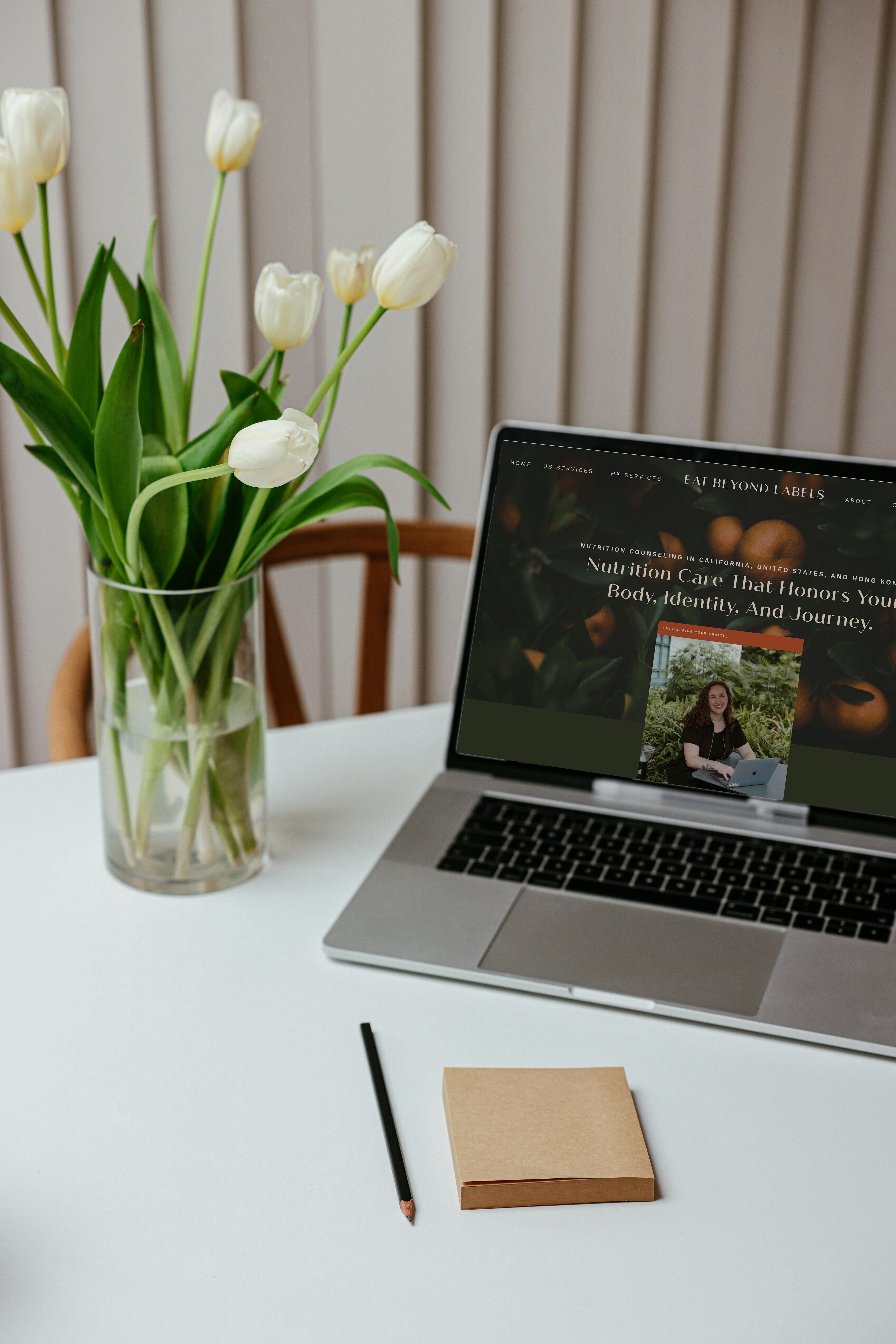 White tulips in a glass vase sit beside an open laptop showing a website, with a notebook and pencil on a white desk.