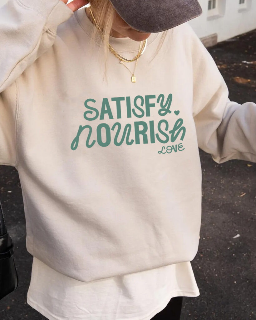 Person in a beige sweatshirt and gray cap displays teal “Satisfy Nourish Love” lettering, conveying a relaxed, positive mood.