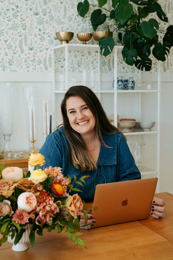 Florist smiling in a denim shirt works on an orange laptop beside a colorful flower arrangement, with plants and patterned wallpaper behind her.