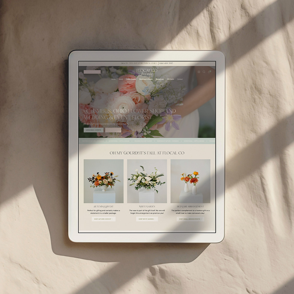 Tablet displaying an elegant floral wedding website with bouquets and cream tones, resting on a textured wall in warm sunlight and shadows.