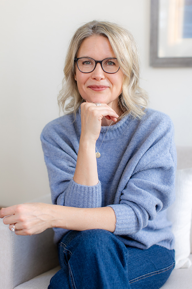 Smiling middle-aged woman with glasses sits casually in a blue sweater and jeans, resting her chin on her hand in a warm, inviting portrait.