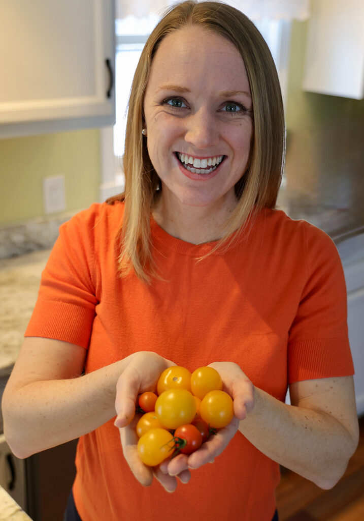 Jen Meijer smiling in an orange shirt holds a colorful handful of cherry tomatoes in a bright kitchen, creating a warm, cheerful mood.