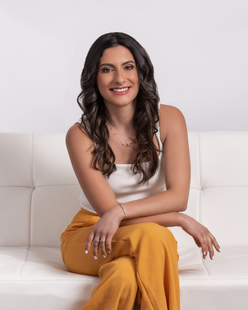 Smiling woman with long, wavy dark hair sits cross-legged on a white sofa, wearing a white top and mustard-yellow pants, conveying warmth.