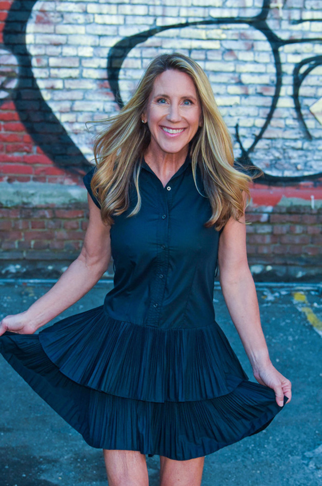 Smiling woman in a sleeveless black dress spreads its pleated skirt against a colorful graffiti-covered brick wall, creating a confident, playful urban portrait.