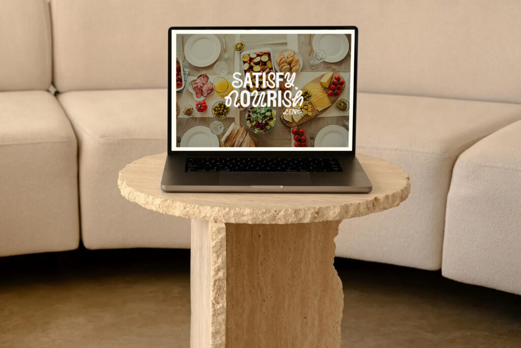 Laptop displaying a colorful food spread with “Satisfy Your Cravings” text, positioned on a stone pedestal table before a beige sofa.