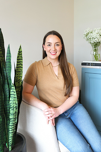 Smiling woman with long brown hair sits in a bright room beside tall snake plants and a blue cabinet topped with white flowers.