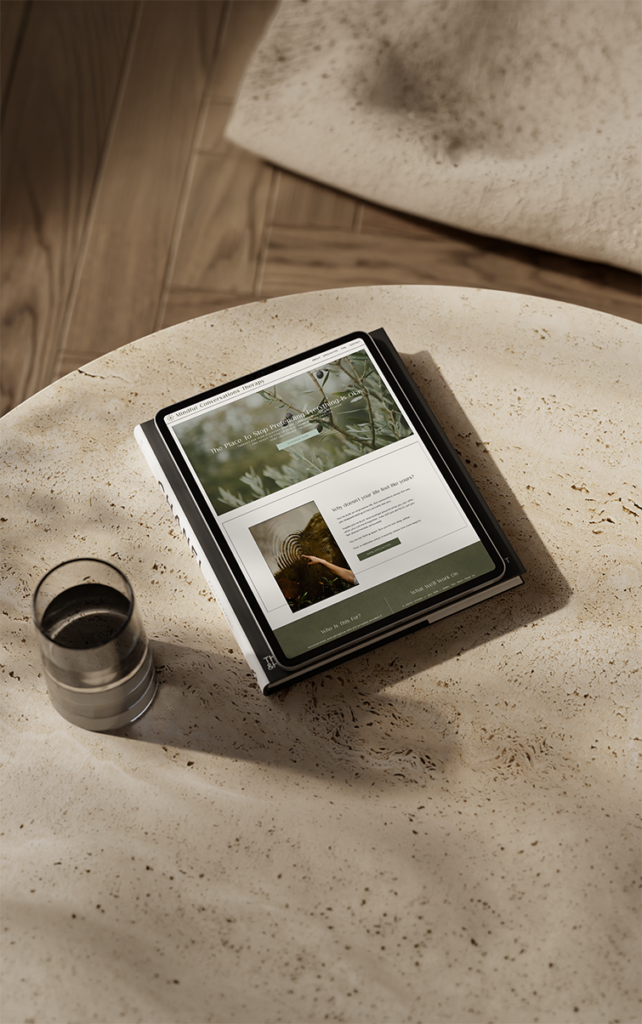 Tablet displaying an earthy therapist website rests on a speckled stone table beside a glass of water, creating a warm, quiet, rustic scene.