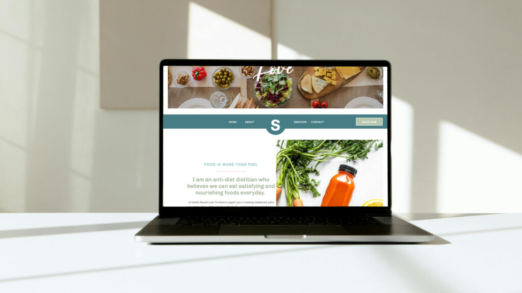 Laptop displaying a colorful food and wellness website with herbs, vegetables, and orange juice, set in a bright, sunlit minimalist room.