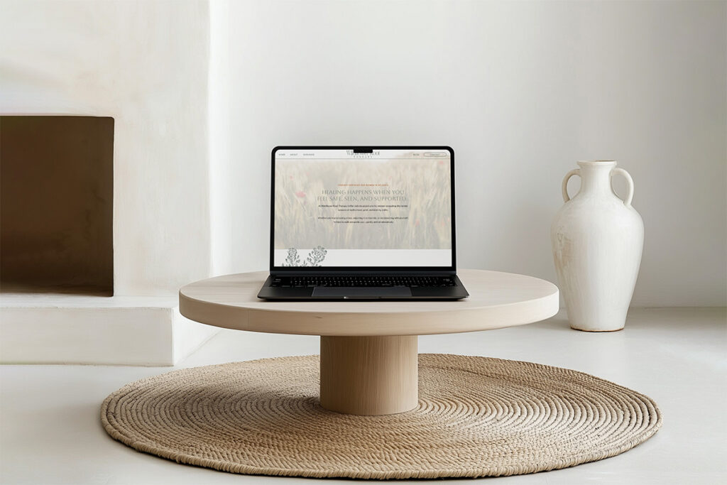Minimalist living room with an open laptop on a round wood table, woven rug below, white vase beside it, and calm neutral decor.