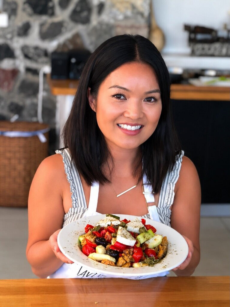 Monique Vuong holds a colorful salad plate at a kitchen table, featuring greens, tomatoes, cucumber, cheese, and olives; warm, inviting mood.