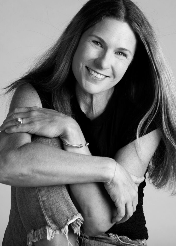 Black-and-white portrait of a smiling woman with long hair, seated and hugging her knees, conveying warmth, confidence, and approachability.
