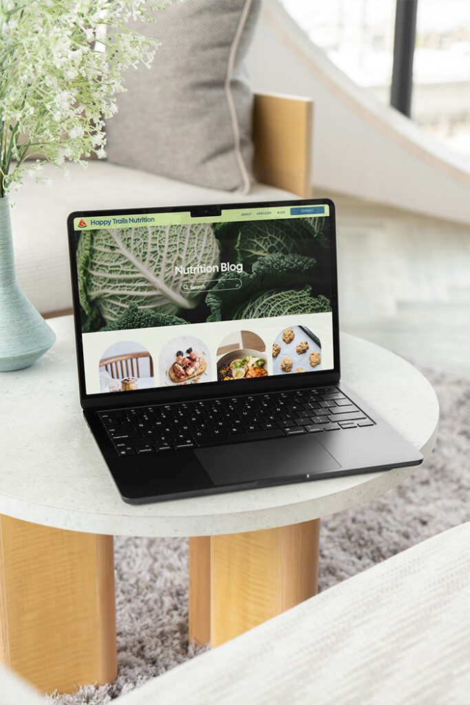 Open laptop displaying a “Nutrition Blog” website with healthy food images, on a round table beside a vase in a bright, cozy room.