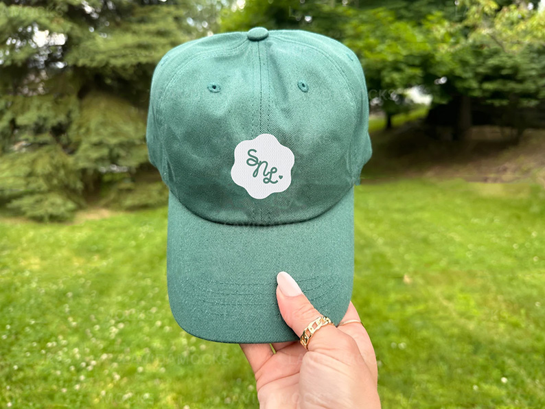 Teal green baseball cap with a white “Snyc” logo patch, held by a hand outdoors against a softly blurred, leafy park background.