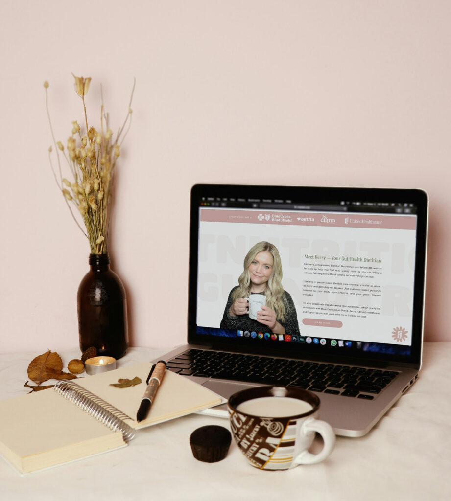 Warm, cozy workspace with a laptop showing a woman’s gut health dietitian website design, notebook and pen, coffee mug, candle, dried flowers, and autumn leaves.