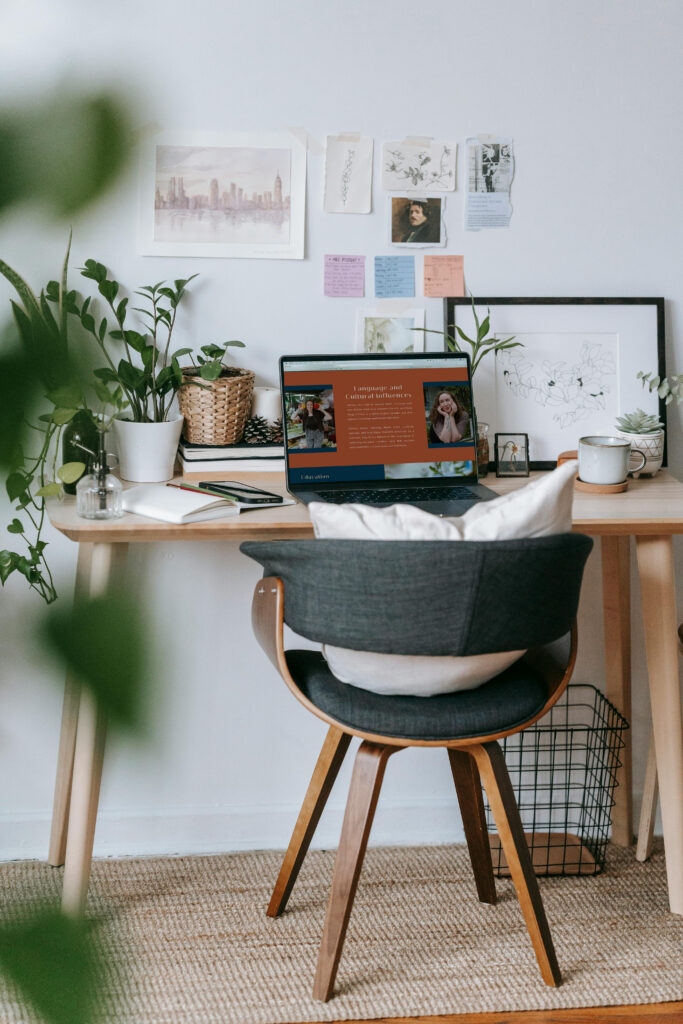 Cozy home office with a laptop on a wooden desk, leafy plants, wall art, and a cushioned chair, creating a calm, creative workspace.