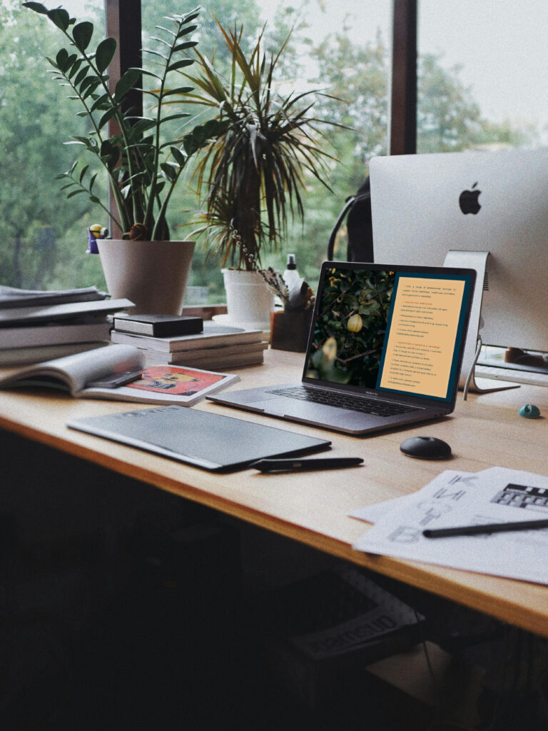 Cozy home office with potted plants, iMac, open laptop displaying notes, notebooks, tablet, and papers on a sunlit wooden desk.