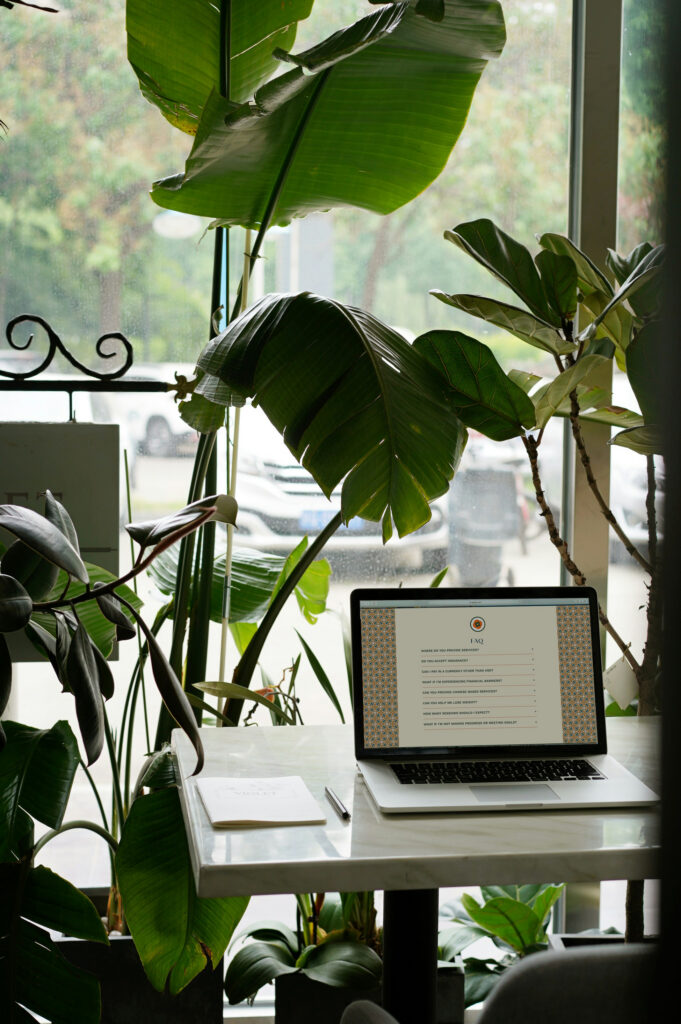 Laptop displaying a document sits on a white desk surrounded by lush tropical plants beside a bright window, creating a calm, green workspace.