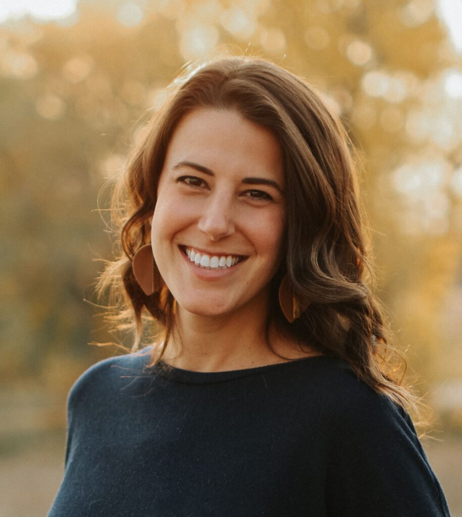 Smiling woman with wavy brown hair and hoop earrings poses outdoors in warm golden-hour light, conveying warmth and confidence.