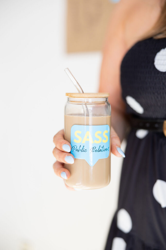 A person in a black dress with white polka dots holds a glass jar of creamy iced coffee with a straw, labeled “SASS Public Relations,” in a bright setting.