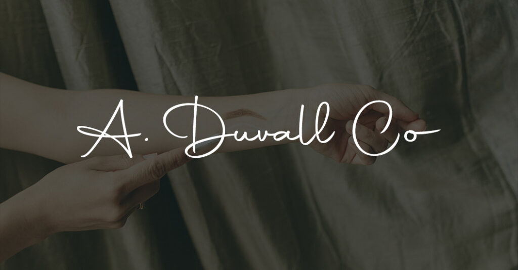 Close-up of hands gently holding dark, textured fabric, overlaid with elegant white script reading “A. Duvall Co.”; calm, refined mood.