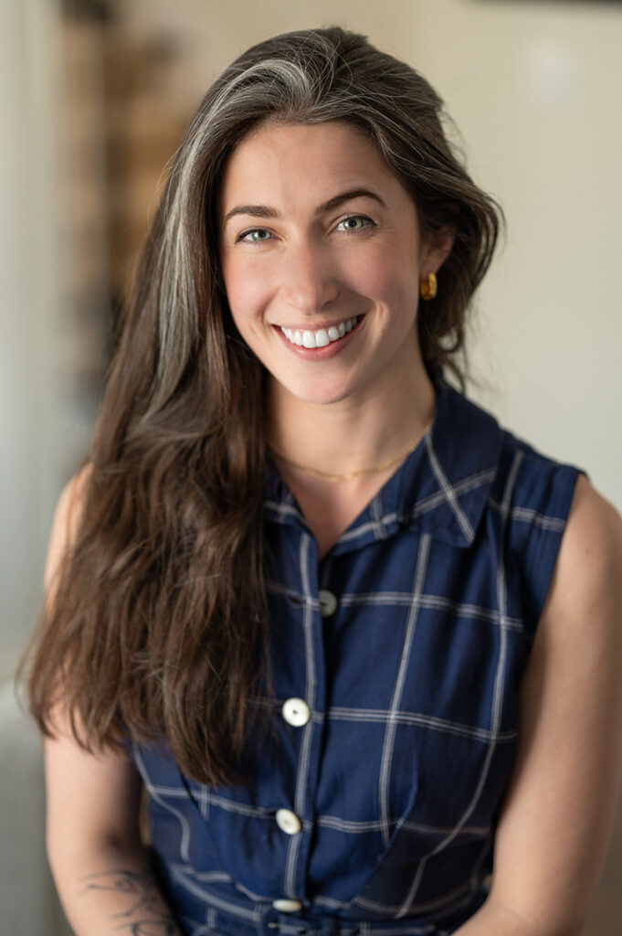 Portrait of a smiling woman with long brown hair wearing a navy plaid sleeveless shirt, conveying warmth and confidence in soft light.