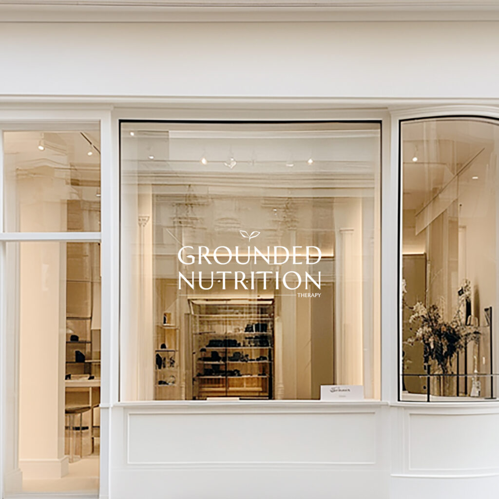 Cream-colored storefront window for Grounded Nutrition Therapy, showing shelves and plants inside; elegant, serene, welcoming wellness atmosphere.