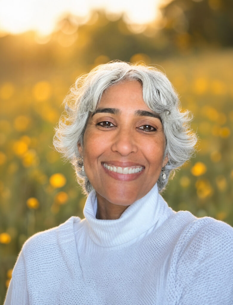 Smiling older woman with short silver hair in a white sweater, posing outdoors amid golden wildflowers in warm, sunlit light.