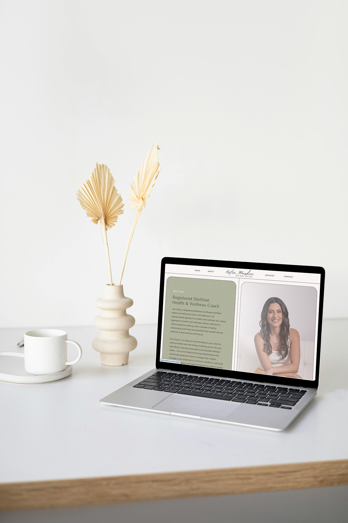 Bright minimalist desk with a laptop displaying a webpage featuring a woman’s portrait and text, beside a coffee cup and dried flowers.