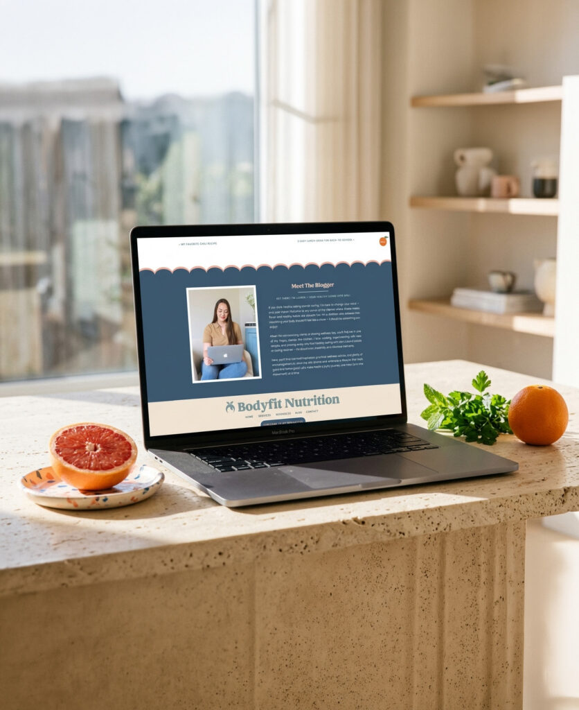Laptop displaying a BodyFit Nutrition webpage sits on a sunlit kitchen counter beside grapefruit, orange, and herbs, creating a fresh, healthy mood.