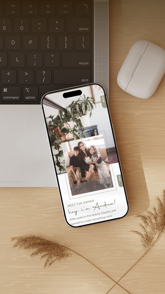 Top-down cozy workspace with laptop keyboard, white mouse, and smartphone showing a stylish family photo collage on a wood desk with pampas grass.