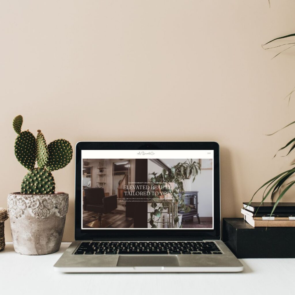 Laptop displaying an interior design website sits on a white desk, framed by a potted prickly pear cactus and leafy plants in a calm, stylish scene.
