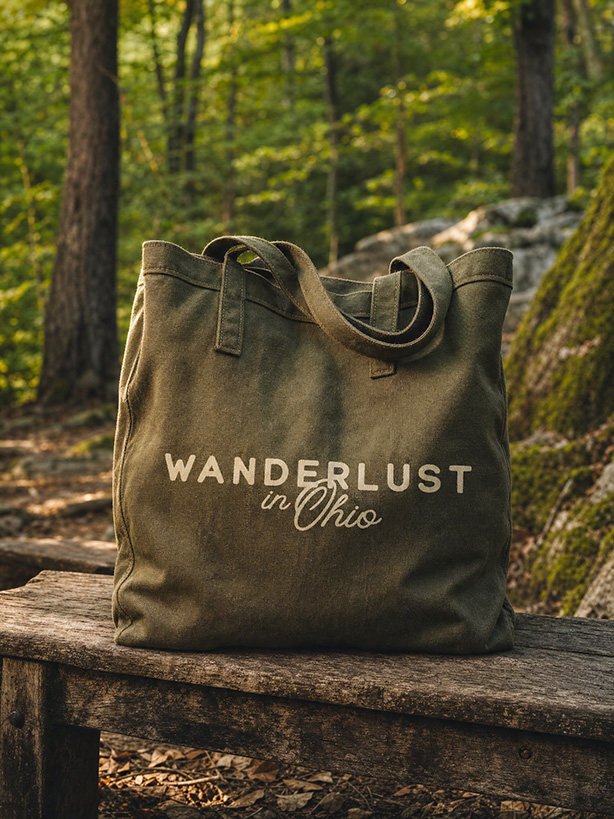 Olive-green canvas tote reading “Wanderlust in Ohio” rests on a weathered log in a sunlit forest, evoking a calm, outdoorsy travel mood.