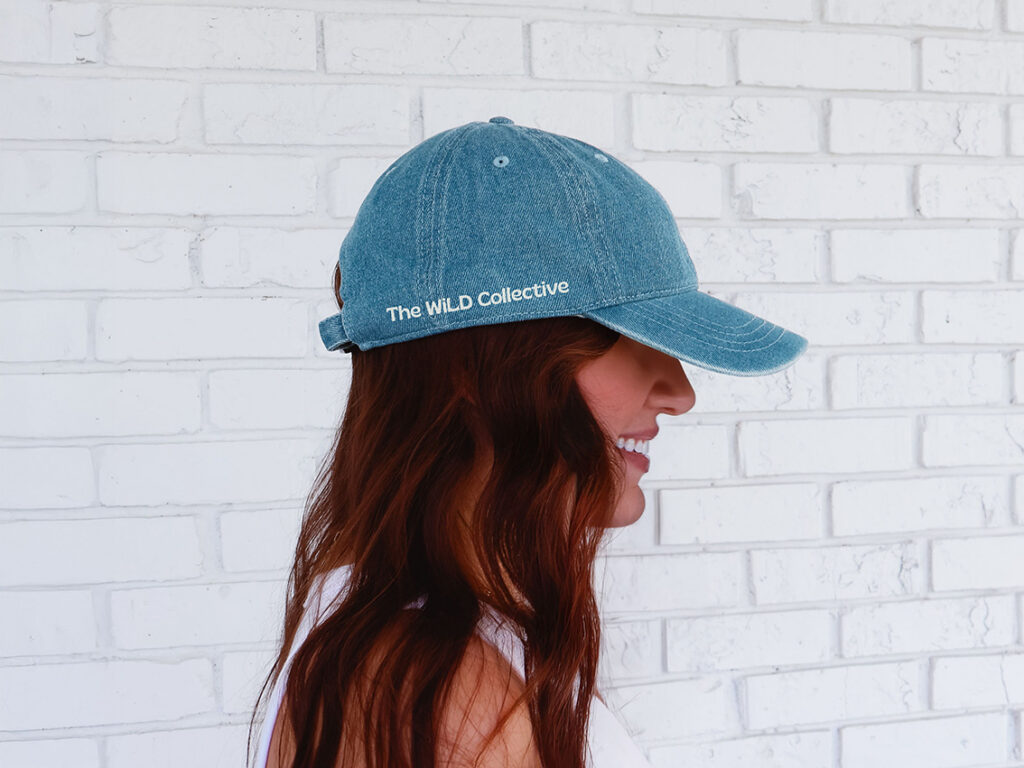 A smiling woman with long auburn hair wears a blue “The WILD Collective” cap, shown in profile against a bright white brick wall.