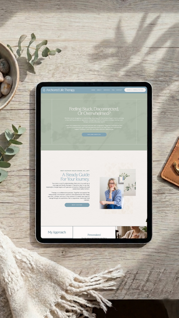 Tablet displaying a mental health website on a sunlit wooden table, surrounded by plants, a blanket, stones, and a leather notebook; calm tone.