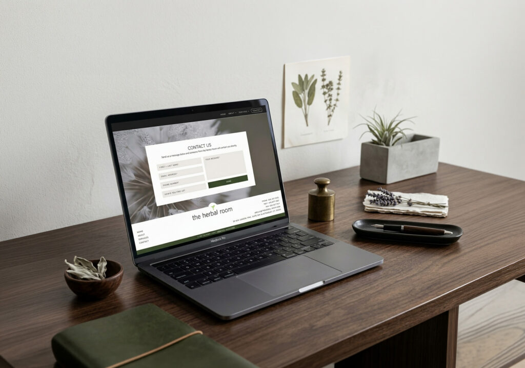 Laptop displaying a clean contact page sits on a wooden desk with a small plant, botanical wall art, and notebook, creating a calm, minimalist workspace.